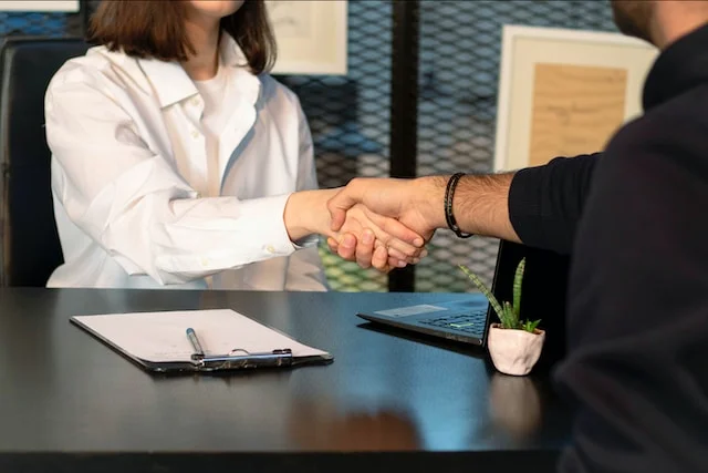 Two people shaking hands across a desk with a clipboard, laptop and plant, closing a deal