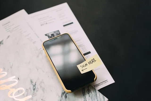 Smartphone resting on a printed contract with a yellow Sign Here sticky note arrow pointing to the screen